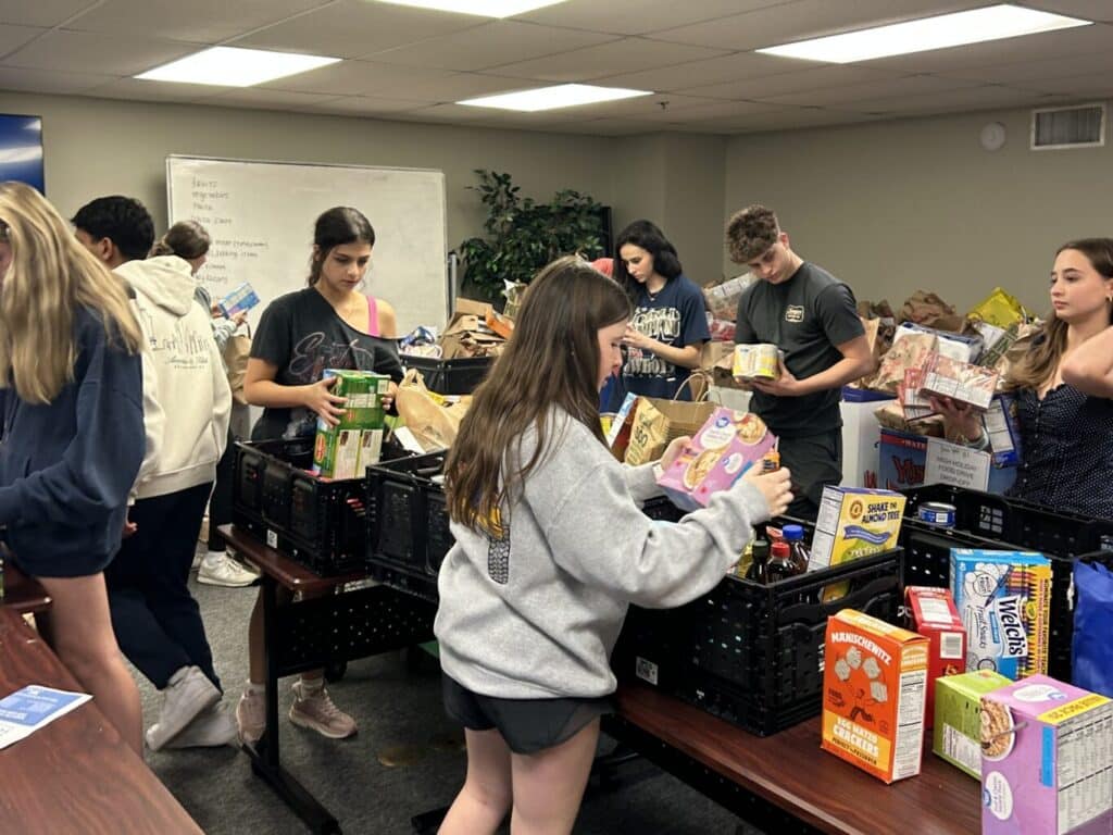 A group of young people sorts donated food items into black crates in a room with tables and a whiteboard.