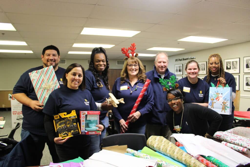 Eight people in matching shirts smile and hold gifts and wrapping paper at a holiday gift-wrapping event in a decorated room.