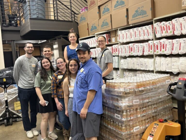 Eight people stand and smile together in a warehouse, surrounded by stacked boxes, canned goods, and bags of rice.