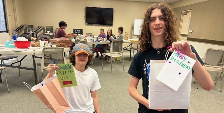 Two teens hold birthday gift bags and cards in a room with people sitting at tables in the background.