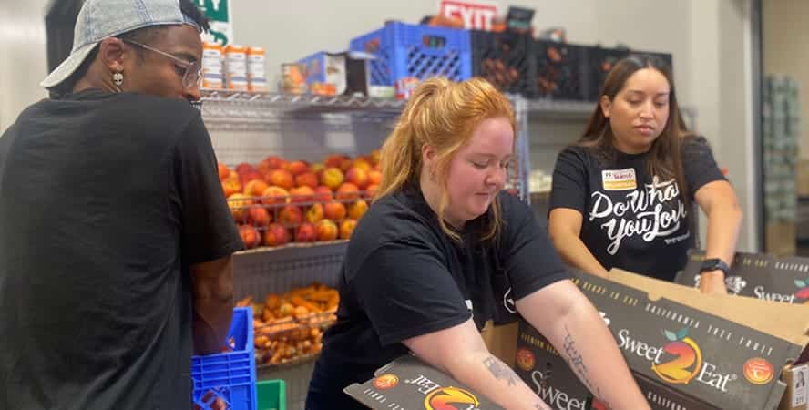 Three people pack fruit into boxes at a food pantry, working together beside shelves of fresh produce.