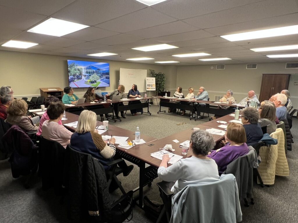 A group of adults sit around tables in a meeting room, listening to a speaker, with papers and drinks in front of them.