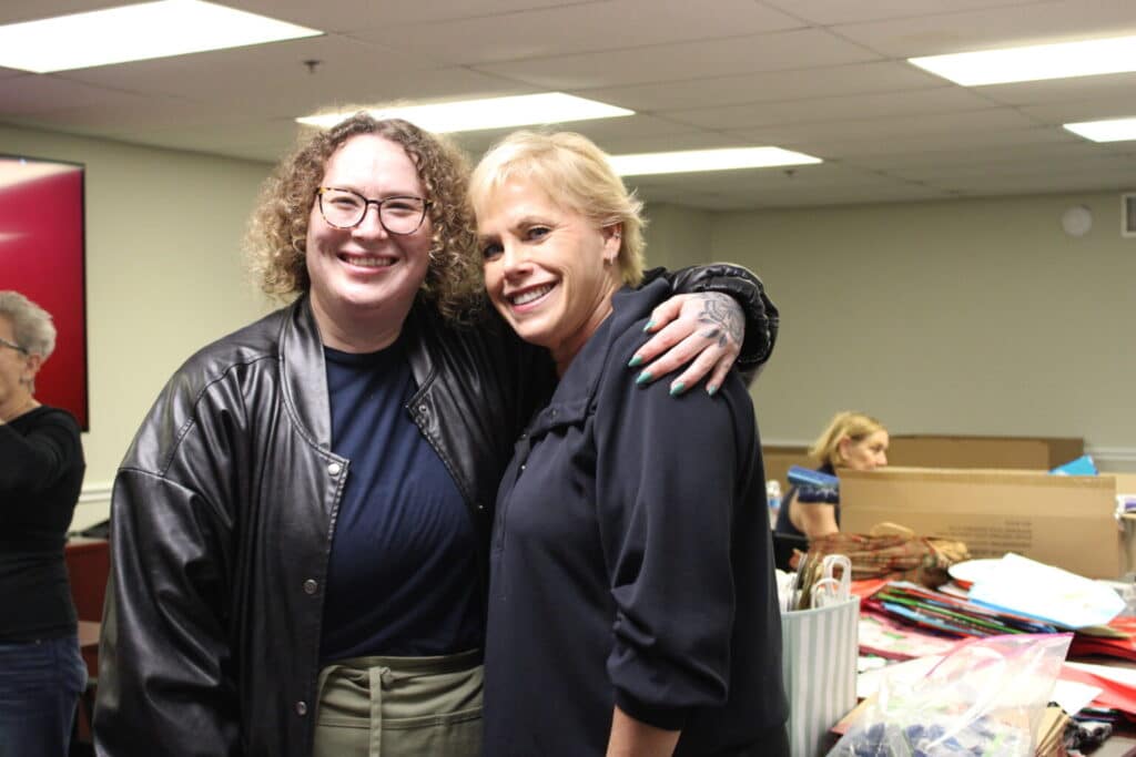 Two people smiling and posing together indoors, with tables and boxes of colorful items in the background.