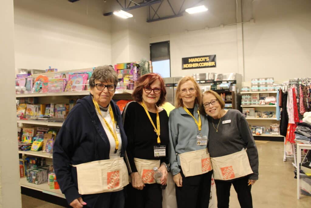 Four women wearing aprons and name badges stand together and smile inside a store with shelves of toys and clothes behind them.