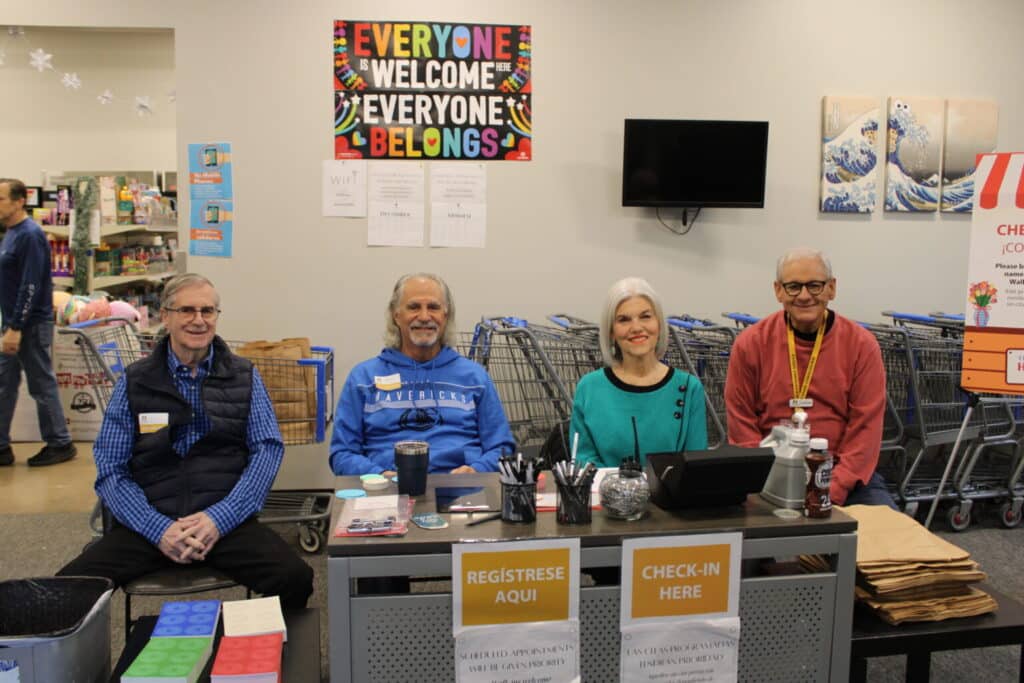 Four smiling adults sit behind a check-in table at an event, with welcome signs and grocery carts in the background.