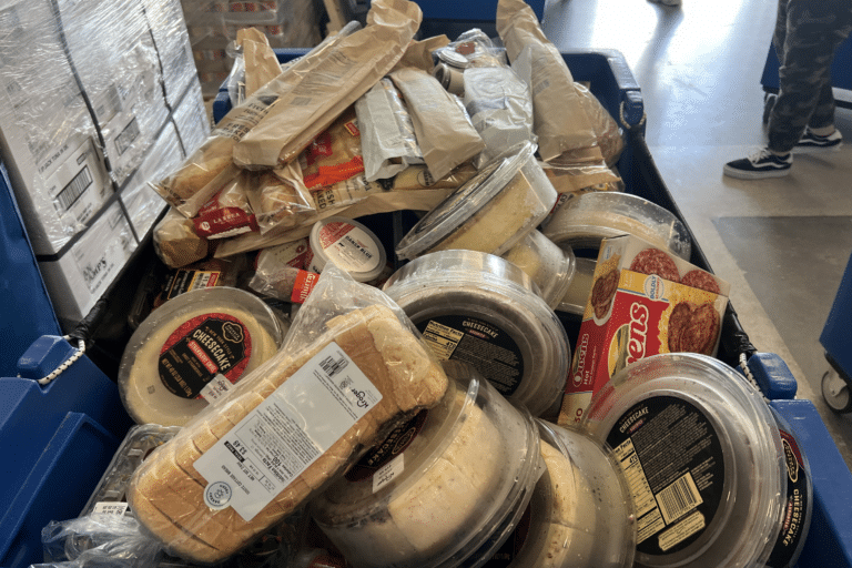 A large blue cart filled with rescued grocery donations, including fresh bread, prepared meals, desserts, dairy products, and packaged foods ready to be sorted at the JFS Dallas Food Pantry.