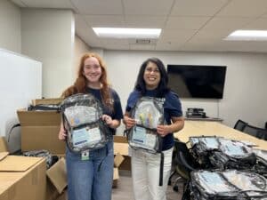 Two JFS Dallas team members hold clear backpacks filled with school supplies as they prepare for the Back-to-School Health Fair.
