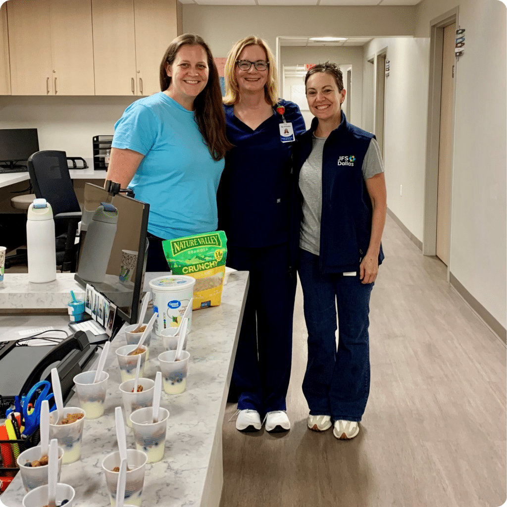 Three JFS Dallas staff members smile together at the JFS Dallas Northpoint Health Center during the Back-to-School Health Fair. Healthy after-school snack samples are displayed on the front desk for families attending the event.