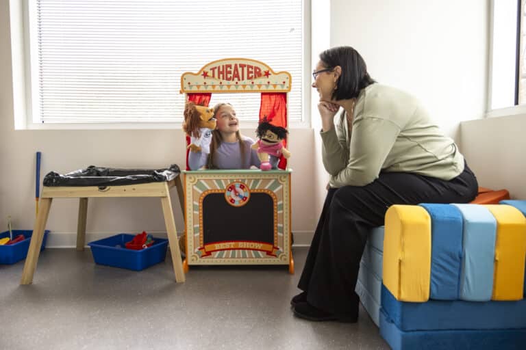 A child puts on a puppet show in a play theater while an adult sits nearby, watching and smiling.