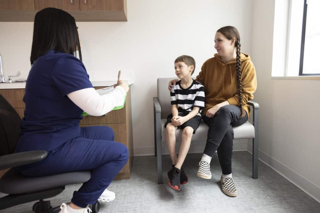 A nurse talks with a young boy and his mother in a clinic room. The boy sits on his mother’s lap.