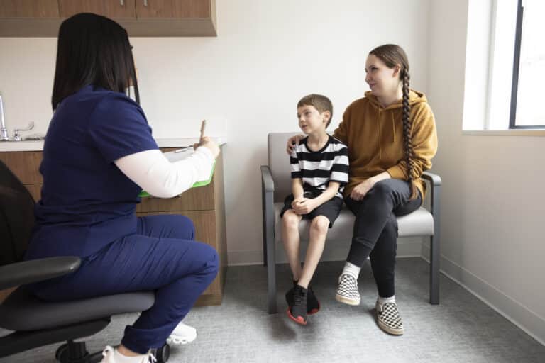 A nurse talks with a young boy and his mother in a clinic room. The boy sits on his mother’s lap.