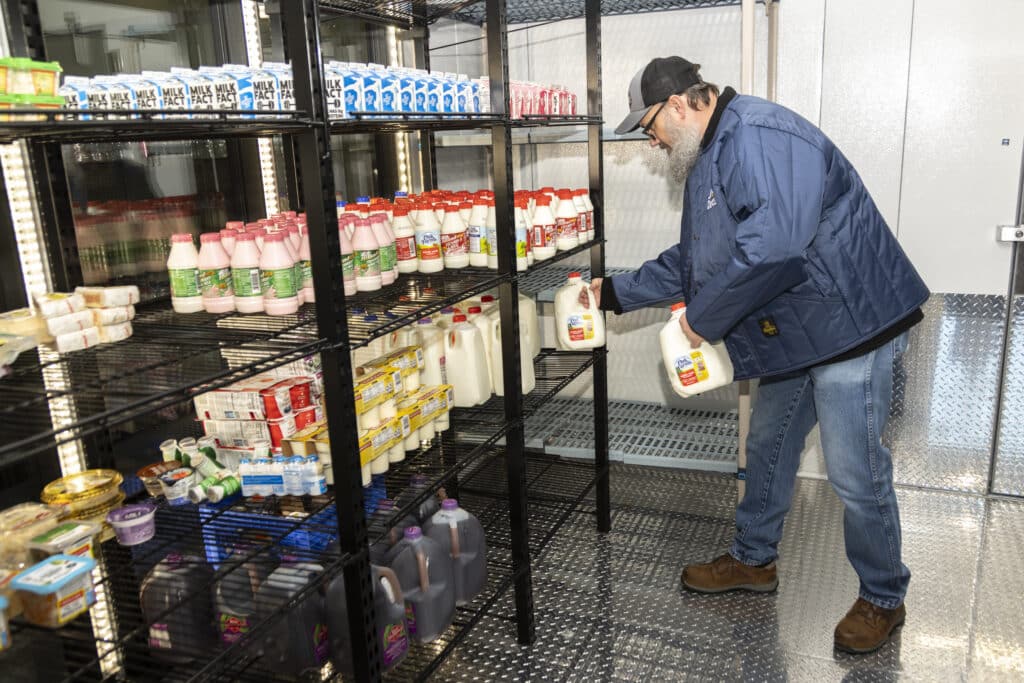 Man stocking milk and other dairy products on metal shelves in a refrigerated storage room.