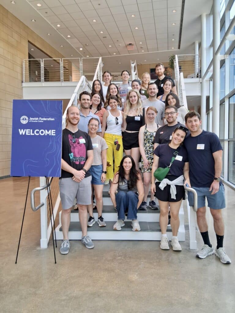 A group of people poses and smiles on indoor stairs beside a “Welcome” sign at a community or organizational event.