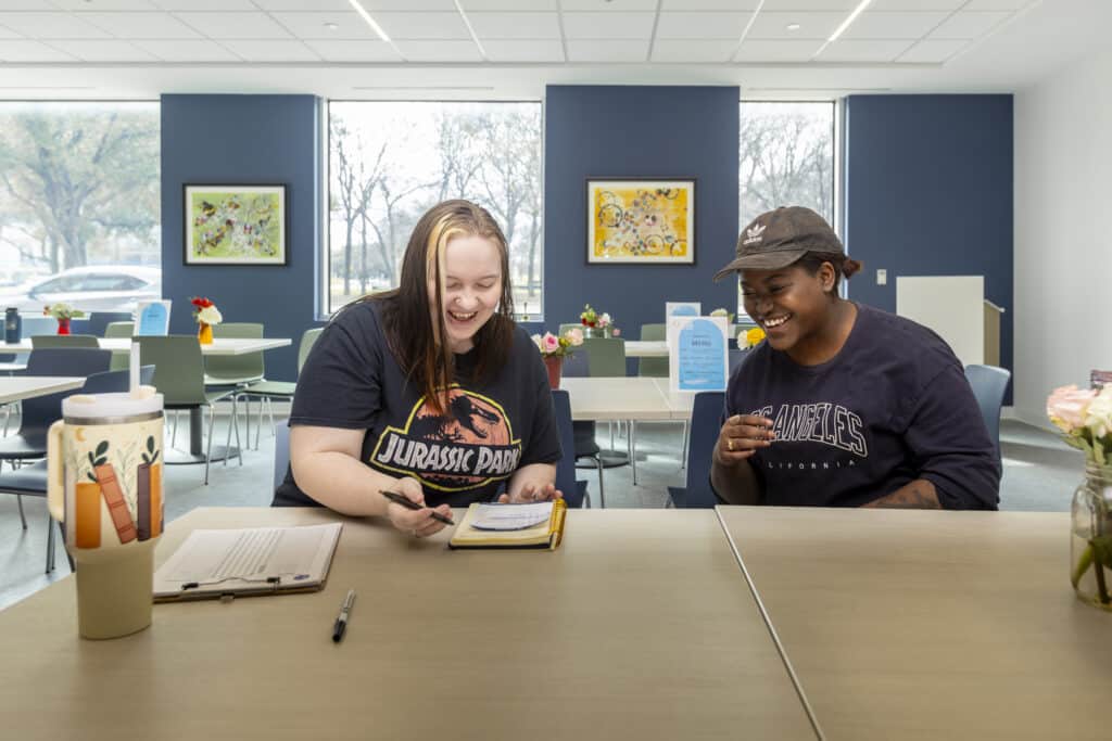 Two women sit at a table, smiling and writing in a notebook in a brightly lit, modern cafe or study area.