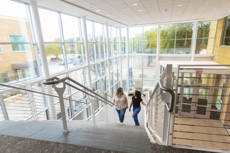 Two women walk up a staircase in a bright, modern building with large windows overlooking a parking lot.