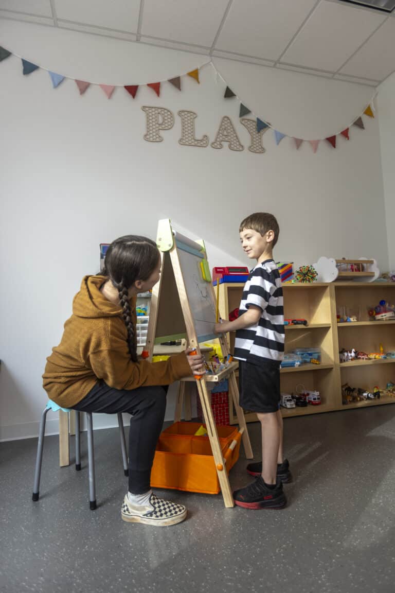 A woman and a boy draw together on an easel in a playroom decorated with toys and a “PLAY” sign.