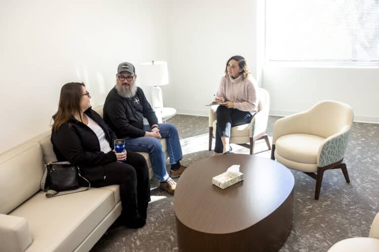 Three people sit in a bright office, two on a couch and one in a chair, talking and taking notes.