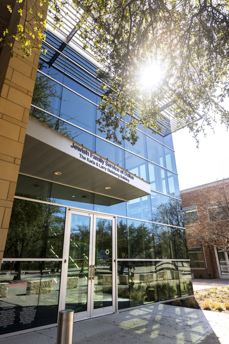 Sun shines on the glass front of Jewish Family Service of Dallas building, reflecting trees and sky.