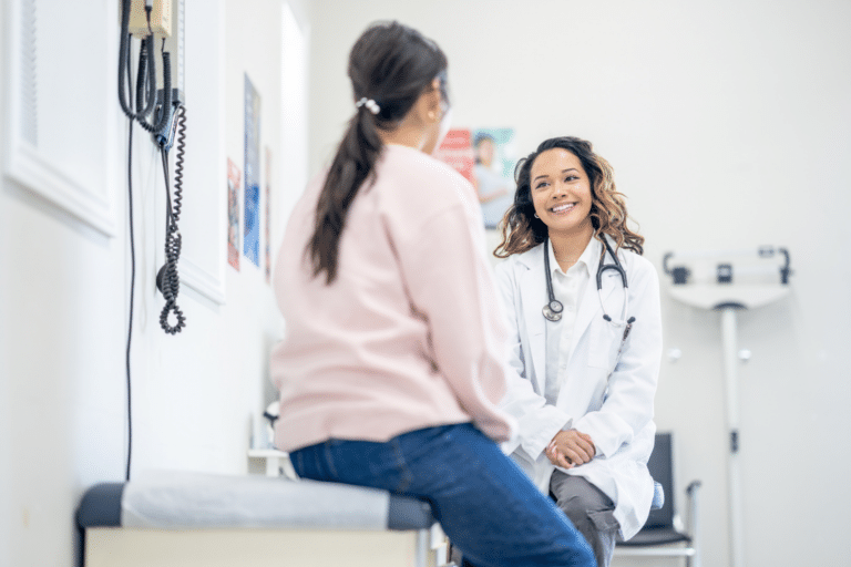 A doctor smiles while talking to a patient sitting on an exam table in a medical office.