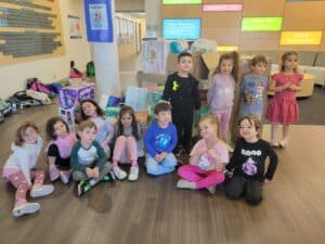 Thirteen young children smile and pose indoors in front of stacked diaper packages and colorful wall signs.