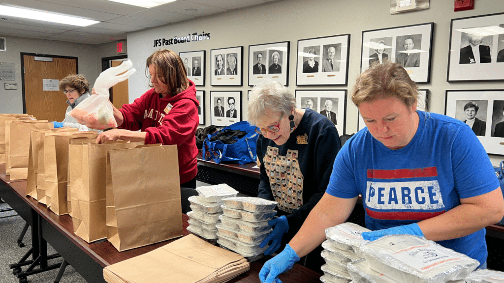 Four women pack food and paper bags on a long table in a room with framed photos on the wall.