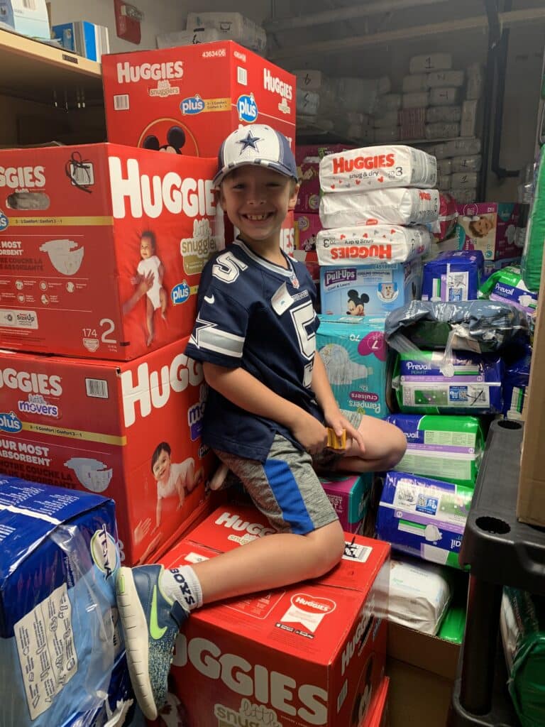 Smiling child in a sports jersey and cap sits on stacked boxes of diapers in a storage room filled with baby supplies.